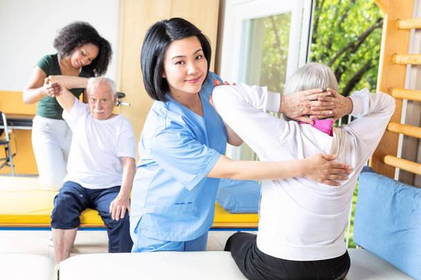 Aziatische en Afrikaanse verpleegkundigen helpen ouderling paar in rehab faciliteit sportschool. - Foto, afbeelding