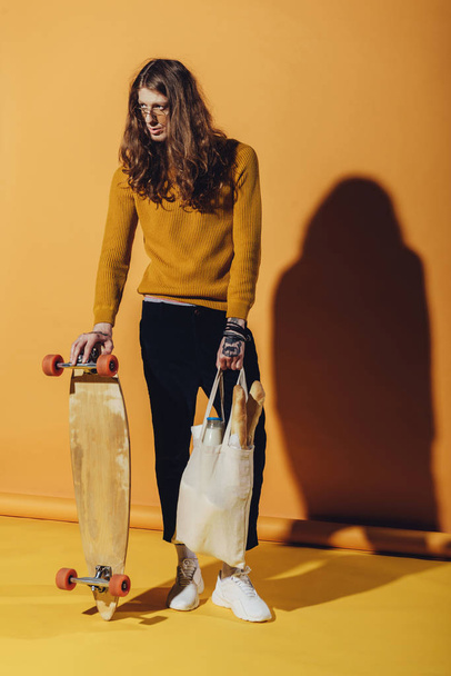 stylish skateboarder holding longboard and bag with food, on yellow  - Foto, afbeelding