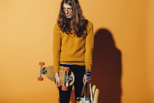 knappe schaatser vasthouden longboard en zak met voedsel, geel  - Foto, afbeelding