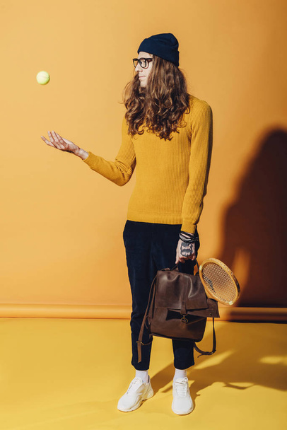 fashionable man with leather backpack and wooden racket throwing up tennis ball, on yellow - Foto, afbeelding