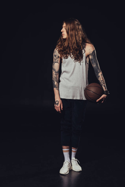 Tattooed man met lange haren die houden van basketbal bal, op zwart - Foto, afbeelding