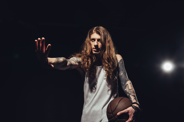 tattooed man with long hair gesturing and holding basketball ball, isolated on black with backlit - Фото, изображение
