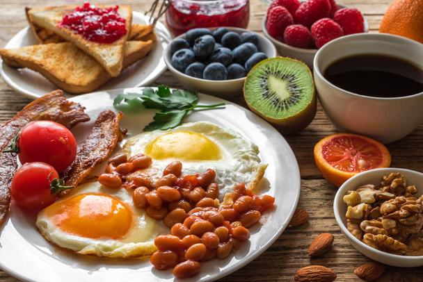 English breakfast. fried egg, beans, tomatoes, coffee, bacon and toast with nuts, fresh fruits and berries - Foto, Bild