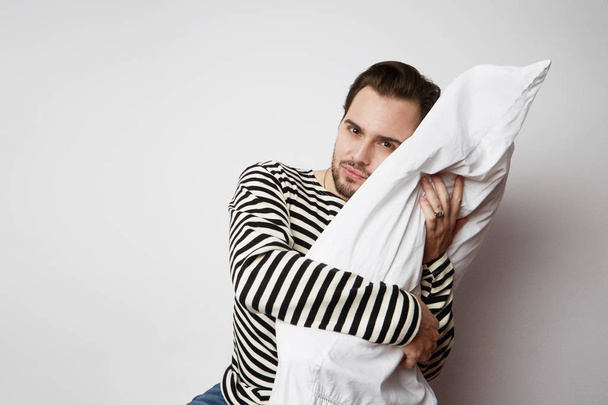 Young pensive man with white soft pillow on empty background. Studio shoot. Horizontal. - Foto, afbeelding