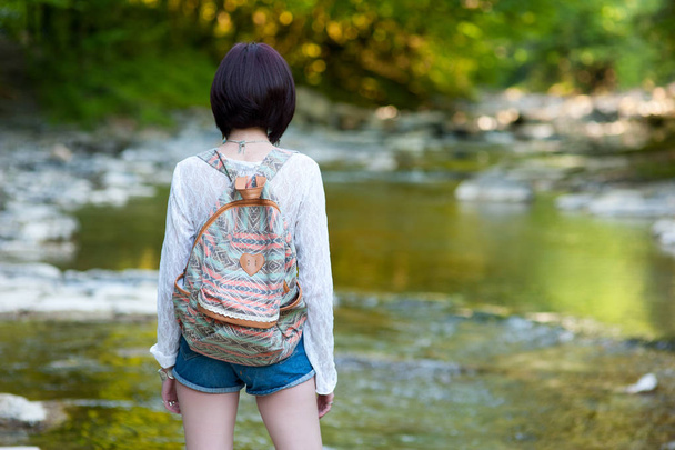A hippie girl in clothes in the style of boho chic and with a backpack is standing with her back turned to the mountain river - Photo, Image