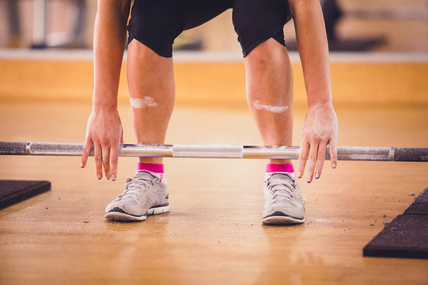 athlete in sneakers and shorts takes a bar with weights by the hands from the floor in the gym - Photo, Image