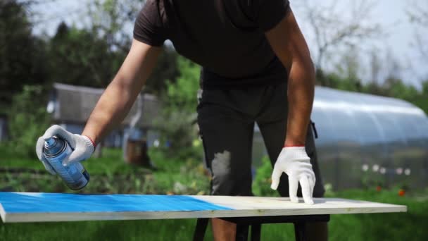 Чоловік рука в робочій рукавичці розбризкує фарбу з розпилювачем на дерев'яну поверхню на відкритому повітрі, чоловік малює меблі своїми руками в саду
 - Кадри, відео