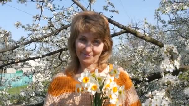 Muttertag. Porträt einer schönen blauäugigen Frau mittleren Alters, die fröhlich lächelnd in die Kamera blickt und vor dem Hintergrund eines üppig blühenden Baumes im Frühling den Duft von Blumen atmet - Filmmaterial, Video