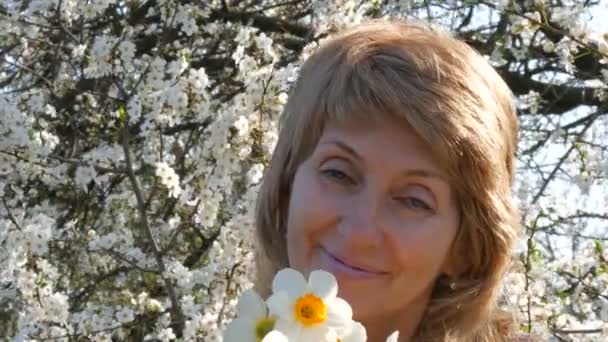 Portret kobiety piękne niebieskie oczy średnim wieku, którzy szczęśliwie patrząc na kamery, uśmiechnięty, oddycha zapach kwiatów na tle zadbanym kwitnące drzewa na wiosnę. Dzień matki - Materiał filmowy, wideo