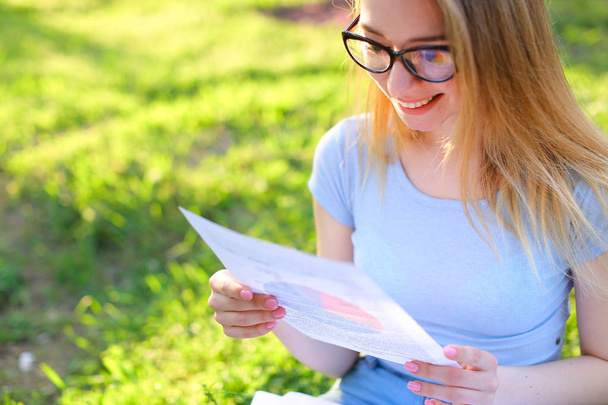 Jovem estudante em óculos de leitura de papéis e sentado na grama verde no parque
. - Foto, Imagem