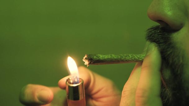 a young guy smoking a rolled jointer with weed buds inside in the green light close-up. - Photo, Image