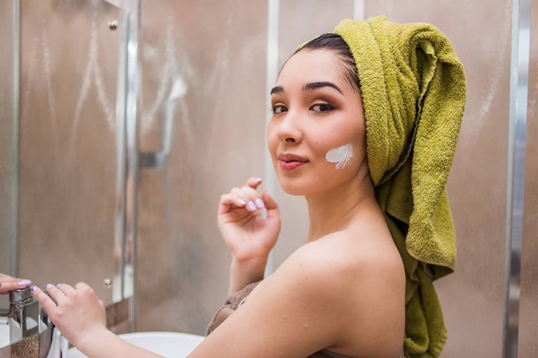 Portrait of a young brunette woman applying cream or face mask in the bathroom - 写真・画像