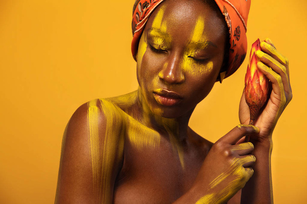 Cheerful young african woman with yellow makeup on her eyes. Female model against yellow background with orange flower. - Фото, изображение