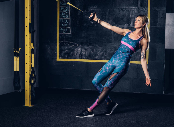 schöne muskulöse blonde Frau in Sportbekleidung mit Tätowierung auf dem Arm, die im Fitnessstudio mit dem Trx-System trainiert. trx-Konzept. - Foto, Bild