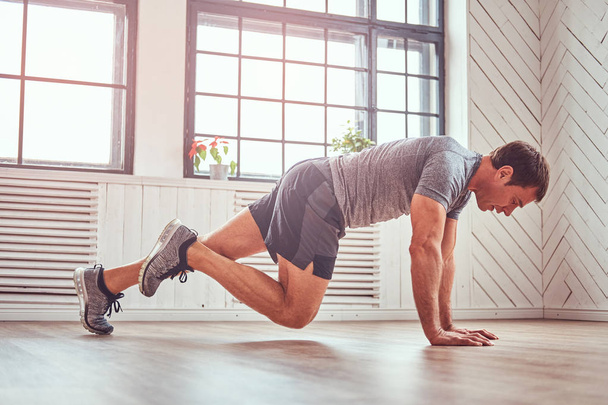 Knappe fitness man in een t-shirt en broek doet functionele oefeningen op verdieping thuis. - Foto, afbeelding