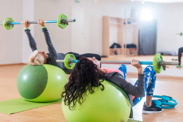 Vista posterior de dos mujeres levantando la barra de pesas tumbadas en la pelota de estabilidad mientras hacen ejercicio en el gimnasio
 - Foto, imagen