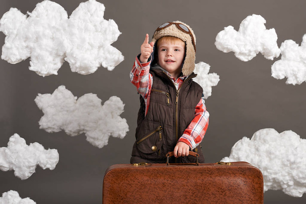 fiú öltözött, mint egy repülőgép kísérleti állni a felhők, a régi bőrönd, és játszik a kézzel készített sík között - Fotó, kép