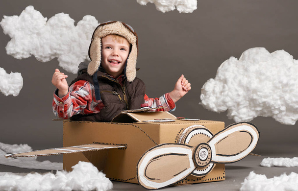 el niño juega en un avión hecho de caja de cartón y sueña con convertirse en piloto, nubes de algodón sobre un fondo gris
 - Foto, Imagen
