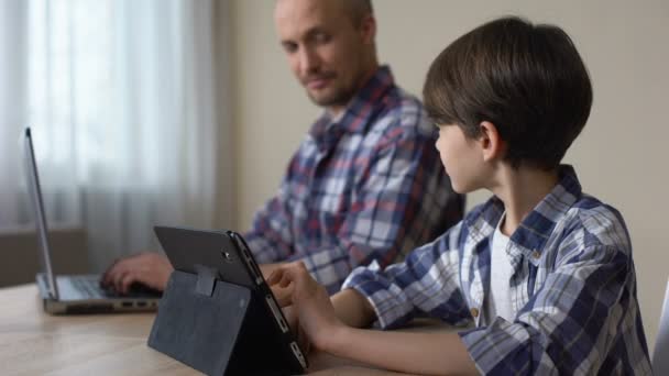 Male kid playing game on tablet while father working on laptop at home, gadget - Video, Çekim