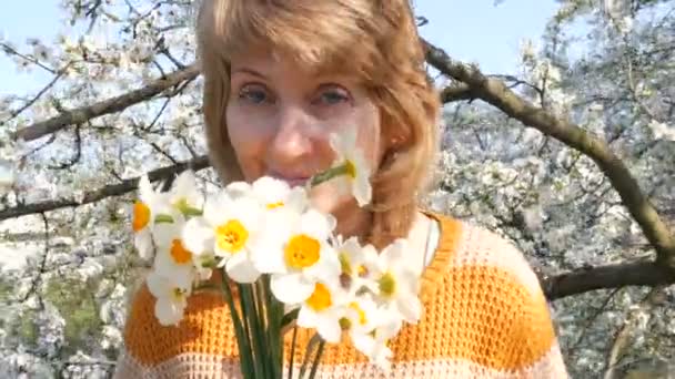 Dia das mães. Retrato de uma bela mulher de meia-idade de olhos azuis que felizmente olhando para a câmera, sorrindo, respira fragrância de flores no fundo de uma árvore florida exuberante na primavera
 - Filmagem, Vídeo
