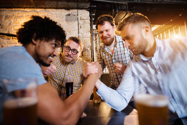 jonge gelukkig bebaarde mannen drinken met bier in de kroeg en met arm worstelen uitdaging  - Foto, afbeelding