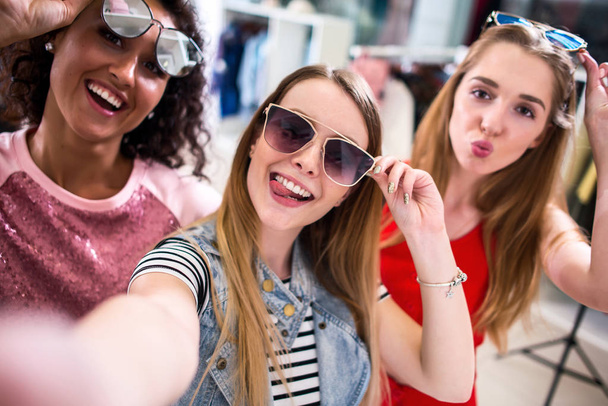 Smiling girlfriends wearing stylish sunglasses having fun time taking selfie with mobile phone while doing shopping in clothing store - Foto, Imagen