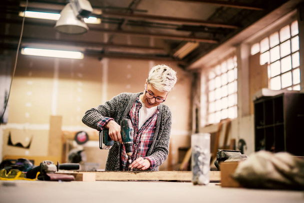 молодая работящая профессиональная женщина плотник с электрическим креслом сверла в мастерской
 - Фото, изображение