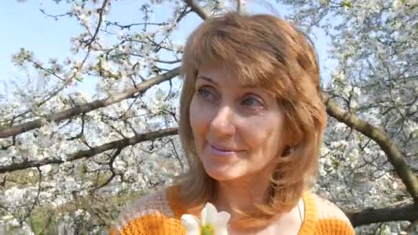 Día de las madres. Retrato de una hermosa mujer de mediana edad de ojos azules que felizmente mirando a la cámara, sonriendo, respira fragancia de flores en el fondo de un árbol floreciente en la primavera
 - Imágenes, Vídeo