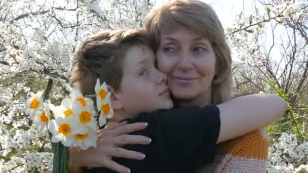 Взрослая мать среднего возраста обнимает и нежно целует своего сына-подростка на фоне красивого роскошного дерева. День матери весной
 - Кадры, видео