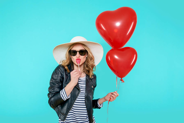 Beautiful rocker girl in leather jacket and summer hat kissing heart shaped lolipop and holding heart shaped balloons. Attractive cool young woman fashion portrait over pastel blue background. - 写真・画像