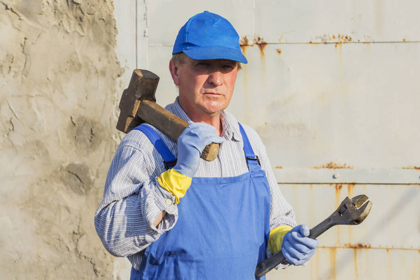 worker in blue overalls with a sledgehammer and a wrench. Serious look. - Photo, Image