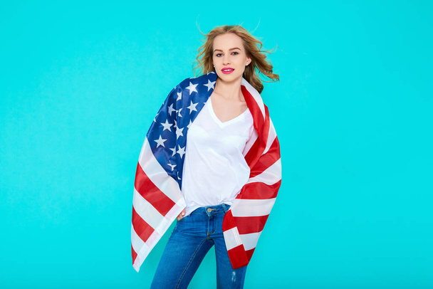 I love America. Happy young smiling woman in jeans and white Tshirt holding American flag and looking at camera while standing against pastel blue background. - Foto, imagen