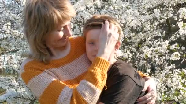 Een volwassen middelbare leeftijd moeder knuffels en kusjes van haar tieners zoon teder tegen de achtergrond van een prachtige weelderige boom. Moederdag in het voorjaar - Video