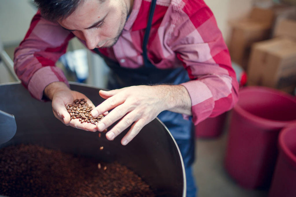 Photo of young man with coffee beans in hand next to roaster - Fotó, kép