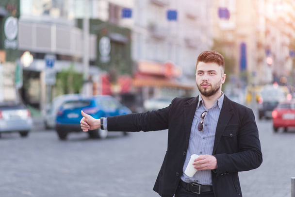 Крепкий бородатый мужчина в деловом костюме стоит у дороги с чашкой кофе в руках и ловит такси. Занятый бизнесмен ловит машину на дороге
 - Фото, изображение