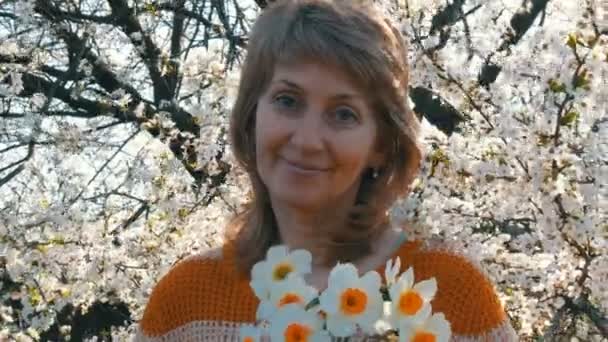 Día de las madres. Retrato de una hermosa mujer de mediana edad de ojos azules que felizmente mirando a la cámara, sonriendo, respira fragancia de flores en el fondo de un árbol floreciente en la primavera
 - Metraje, vídeo