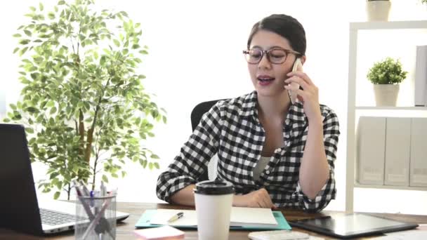 professional young female office worker using mobile cell phone calling for friend chatting when she finished work at home sitting on workplace looking at window relaxing. - Video, Çekim