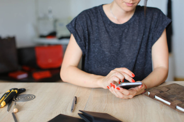European female person looking for ideas by smartphone near handmade notebook at leather atelier. - Фото, изображение