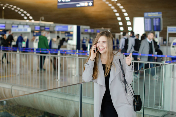 Perosn feminino bonito falando por smartphone no corredor do aeroporto, vestindo casaco cinza e saco
. - Foto, Imagem