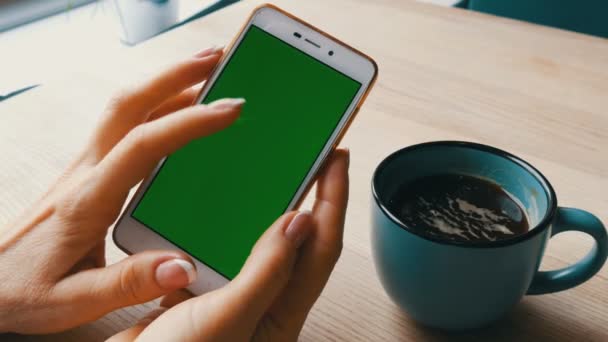 Smartphone de pantalla verde. Chroma Key en un teléfono inteligente blanco, las manos femeninas sostienen el teléfono móvil en un café al lado de una taza de café
 - Imágenes, Vídeo