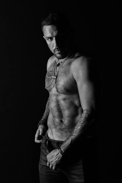 Bearded man with tattooed body. Tattoo model with six pack and ab. Macho with sexy bare torso in jeans. Sportsman with muscular chest and belly. Sport or fitness and bodycare, black and white - Foto, immagini