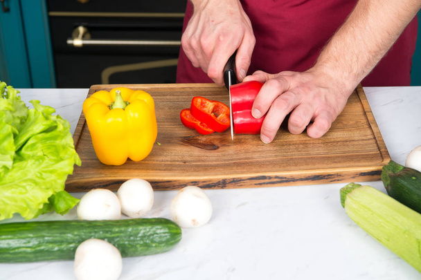 Vegetables getting cut on wooden cutting board. Hand slice pepper with ceramic knife. Food preparation and cooking recipes. Vegetarian menu and healthy diet. Organic products concept - Zdjęcie, obraz