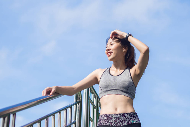 Young woman relaxing after jogging exercise on fence at park to freshen her body and enjoy warm light in morning. Young asian woman take a break from running execise. Outdoor exercise activity. - Foto, Imagem