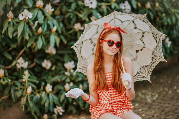 Funny little girl wearing gingham playsuit, heart shaped sunglasses, white gloves, holding lace parasol, retro stylized image - Photo, Image