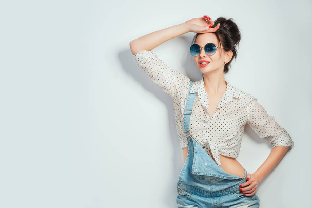 Young beautiful cheerful woman with hair bun in sunglasses wearing in jeans shorts dancing and posing over white wall. Good mood. Hands up! People Emotions Beauty Fashion Lifestyle concepts - Фото, изображение