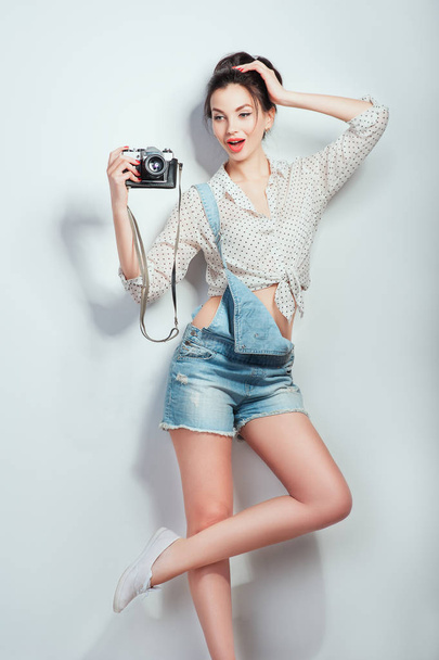 Mode-Look, ziemlich coole junge Frau Modell mit Retro-Kamera tragen in Jeans Kleidung posiert auf weißen Wand. ausdrucksstarke Schönheit Mädchen Fotografin hält Fotokamera. Emotionen Lebensstil Menschen Konzepte - Foto, Bild