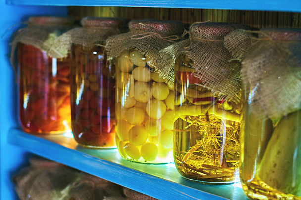 Glass jars covered with burlap fabric with different homemade canned food, preserved vegetables on wooden shelves - Φωτογραφία, εικόνα