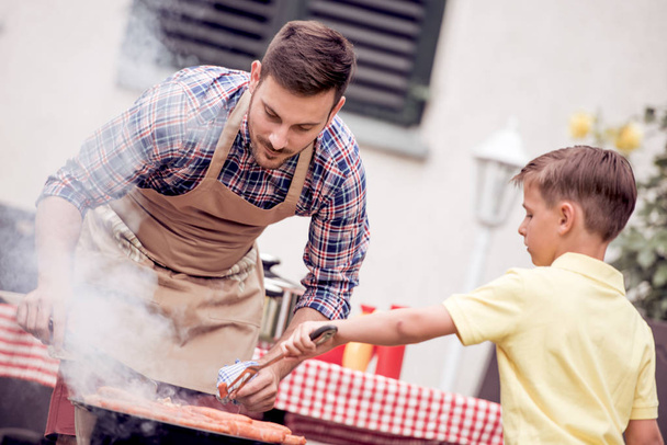 Conceito de lazer, comida, pessoas e feriados - homem que cozinha carne em churrasqueira com seu filho, para sua família na festa ao ar livre de verão
. - Foto, Imagem
