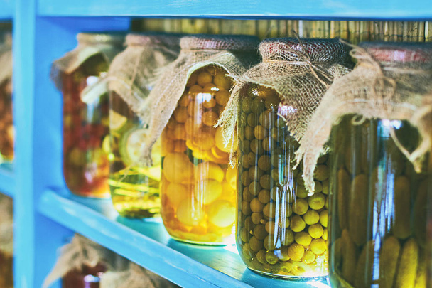 Glass jars covered with burlap fabric with different homemade canned food, preserved vegetables on wooden shelves - Foto, afbeelding