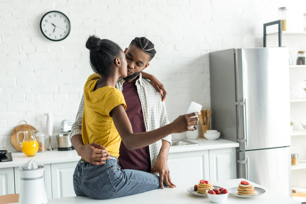 coppia afroamericana baciare e scattare selfie con smartphone in cucina
 - Foto, immagini